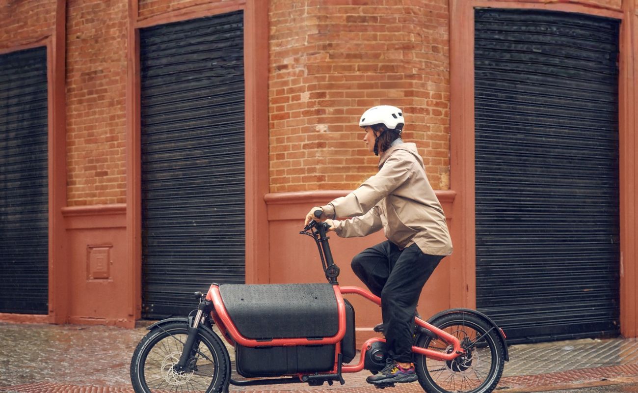 Helmeted rider on a Coral Riese & Müller Carrie2 FAMILY electric cargo bike with front cargo box on a wet street