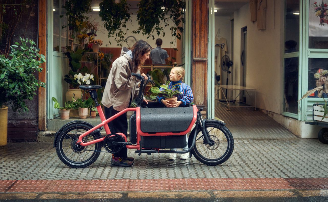Adult and child with a potted plant beside a Coral Riese & Müller Carrie2 FAMILY electric cargo bike outside a shop
