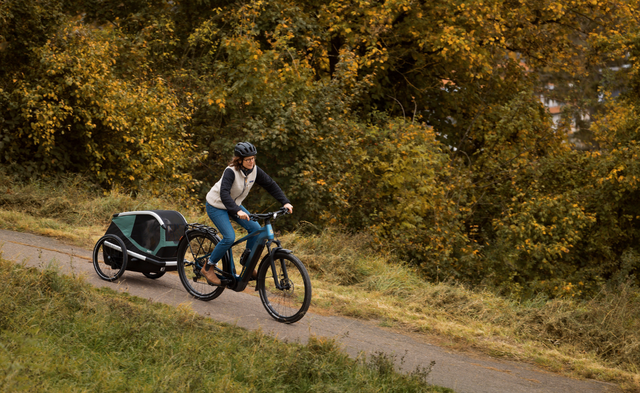 Person riding a Focus F26 Aventura2 6.8 800Wh E-Bike with a child in a trailer on a path surrounded by trees.