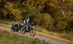 Person riding a Focus F26 Aventura2 6.8 800Wh E-Bike with a child in a trailer on a path surrounded by trees.