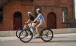 Two people riding Kalkhoff K26 Image L Excite Wave and Gent eBikes on a sidewalk in front of a brick building. | Electric Bikes Brisbane
