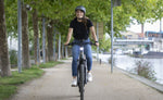 Woman riding a Moustache Open Xroad 4 2026 E-Bike on a path by a river with trees lining the background 