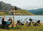 Two people sitting at a picnic table near a lake having carried their picnic on their Orbea Muga electric bikes