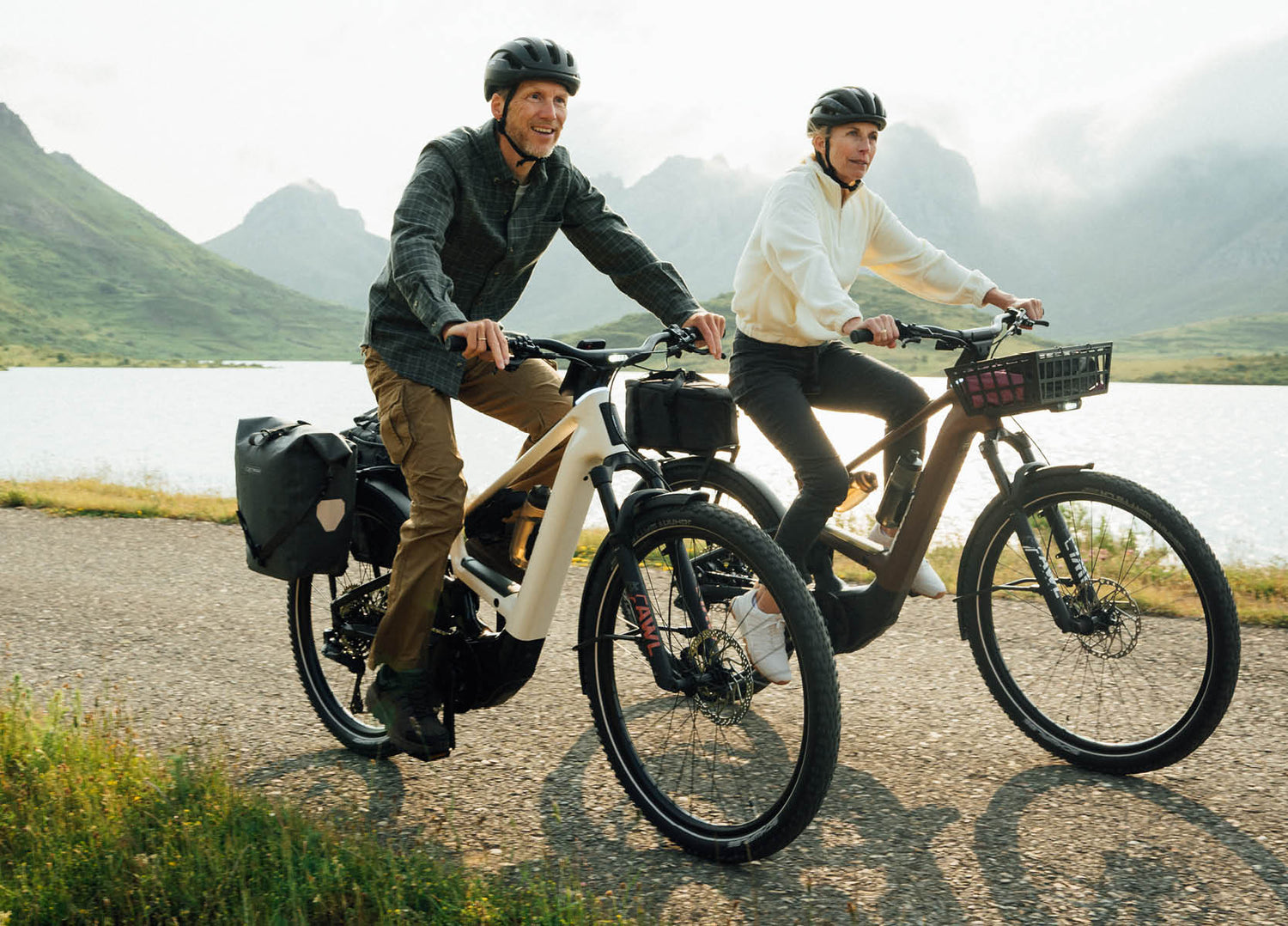 Couple riding Orbea Muga electric bikes on a scenic road with mountains and a lake in the background.