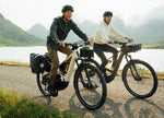 Couple riding Orbea Muga electric bikes on a scenic road with mountains and a lake in the background.