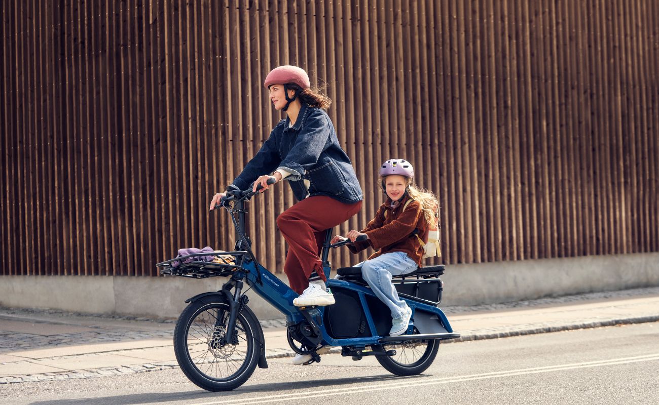 Two people riding a Riese & Muller Multitinker2 Silent Cargo E-Bike on a street with a wooden wall in the background | Electric Bikes Brisbane