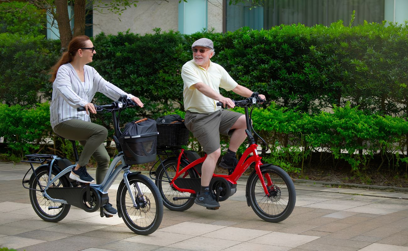 Two adults riding red and grey Tern HSD electric bikes with front baskets along a paved path.