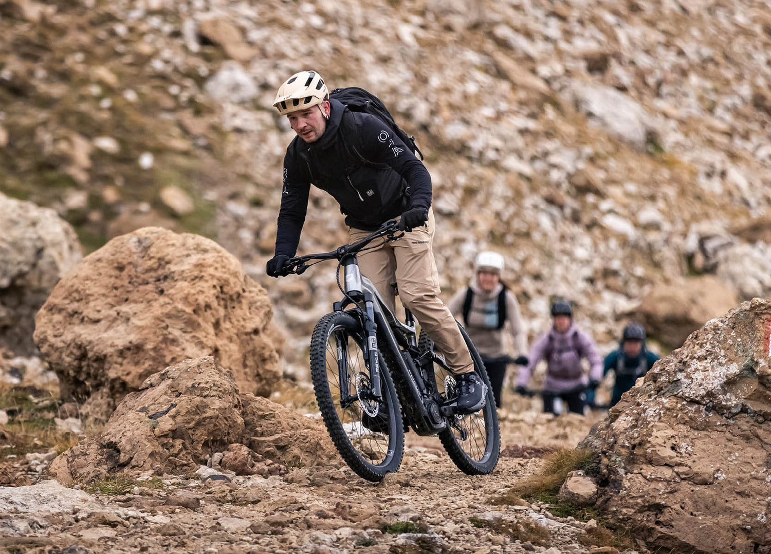 Man riding a F25 Focus Thron2 6.8 ABS electric mountain bike on a rocky path with other riders in the background.