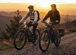 Two people riding equipped Focus Thron2 electric bikes on a mountain trail at sunset. 